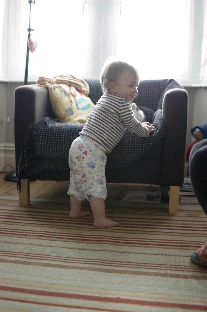 A toddler in owl-print pants pulls herself upright against an armchair in a bright living room — first steps.