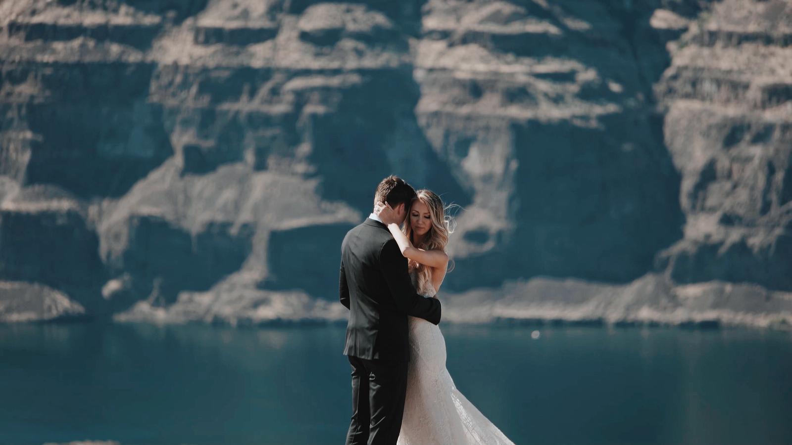 A bride and groom embrace on a cliff edge above deep teal water, her arm around his neck, wind lifting her hair.